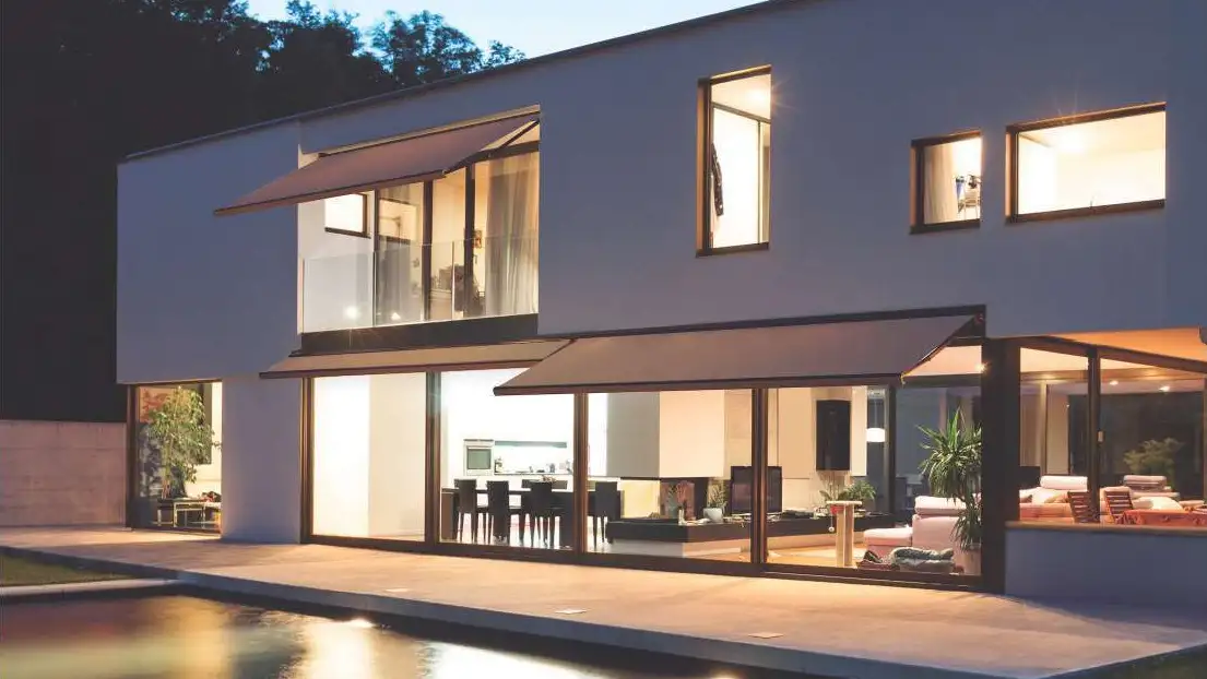 Stores bannes coffre Mini Select 410 à toile brune sur la façade d’une maison contemporaine éclairée de nuit, au bord d’une piscine